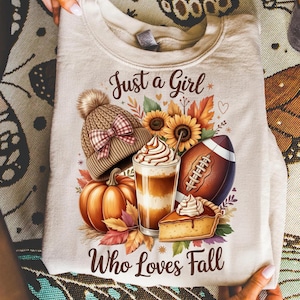 K&ouml;nnte beinhalten: Beiges Sweatshirt mit einem herbstlichen Motiv. Das Design zeigt eine Strickm&uuml;tze, Sonnenblumen, einen Football, einen K&uuml;rbis, einen Latte und ein St&uuml;ck Kuchen. Der Text lautet "Just a Girl Who Loves Fall."