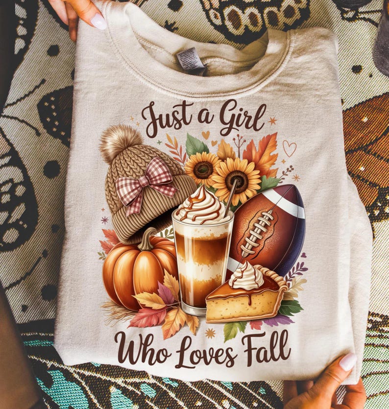 K&ouml;nnte beinhalten: Beiges Sweatshirt mit einem herbstlichen Motiv. Das Design zeigt eine Strickm&uuml;tze, Sonnenblumen, einen Football, einen K&uuml;rbis, einen Latte und ein St&uuml;ck Kuchen. Der Text lautet "Just a Girl Who Loves Fall."