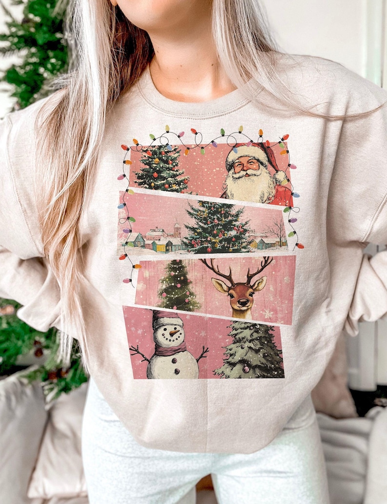 May include: Beige sweatshirt featuring a vintage Christmas design. The design includes Santa Claus, a decorated Christmas tree, a reindeer, and a snowman, all set against a pink background. The design is framed with string lights.