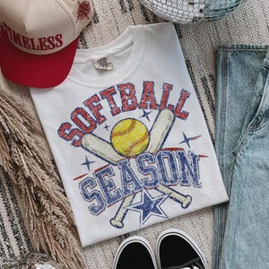 May include: White t-shirt with a distressed "SOFTBALL SEASON" graphic in red and blue, featuring a yellow softball and crossed bats. A red and cream baseball cap with "TIMELESS" text is also visible.