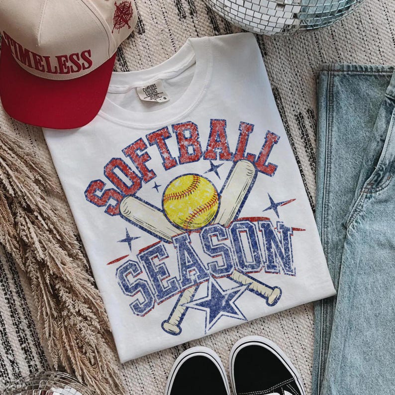 May include: White t-shirt with a distressed "SOFTBALL SEASON" graphic in red and blue, featuring a yellow softball and crossed bats. A red and cream baseball cap with "TIMELESS" text is also visible.