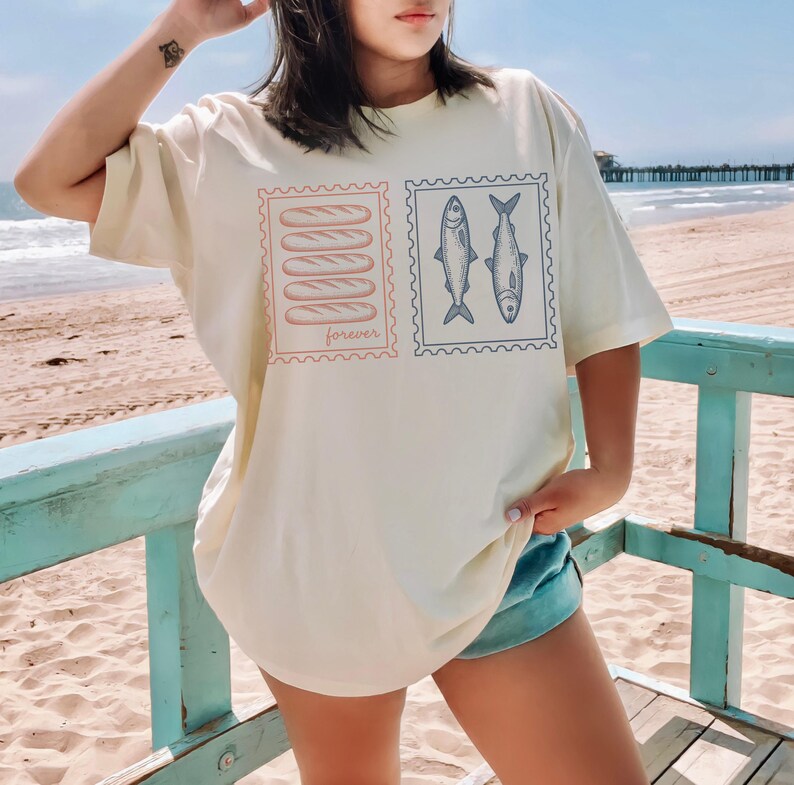 May include: Cream-colored t-shirt featuring two postage stamp designs. One stamp shows five loaves of bread with the word "forever" below. The other stamp displays two fish. The shirt is a relaxed fit.
