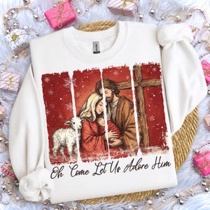 May include: White sweatshirt featuring a religious scene with a family, a lamb, and a cross, set against a red background with snowflakes. The text "Oh Come Let Us Adore Him" is printed at the bottom. A festive holiday design.