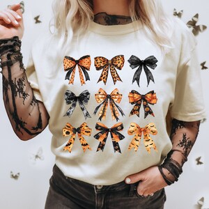 May include: Cream-colored t-shirt featuring a collection of nine watercolor bows in various designs. The bows include orange and black Halloween-themed patterns, leopard print, and solid black. The bows are arranged in three rows of three.