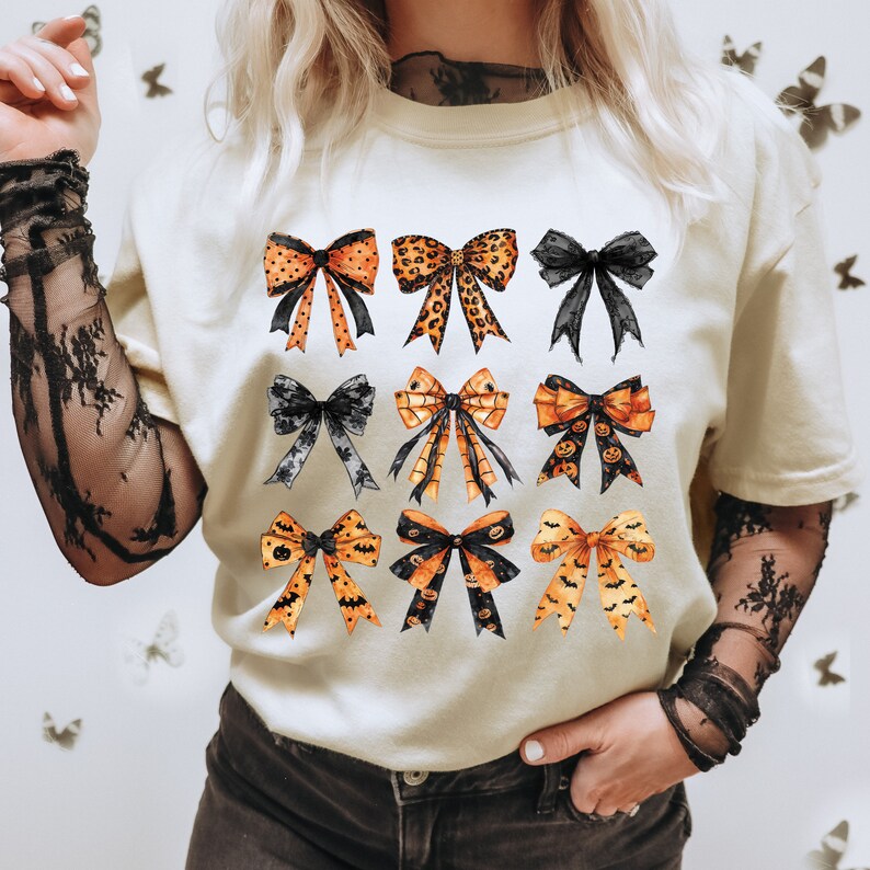 May include: Cream-colored t-shirt featuring a collection of nine watercolor bows in various designs. The bows include orange and black Halloween-themed patterns, leopard print, and solid black. The bows are arranged in three rows of three.