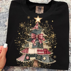 Peut inclure: T-shirt noir avec un motif de sapin de Noël. L'arbre est fait de tissu patchwork aux motifs rouges, verts et crème, orné d'un nœud rouge, de boules et d'une étoile dorée. Des paillettes et des flocons de neige dorés entourent l'arbre.