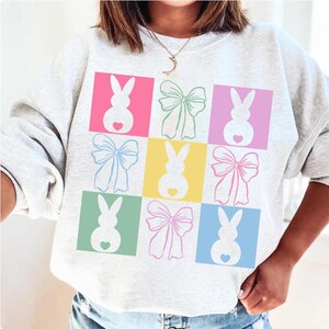 May include: A light gray sweatshirt with a colorful design of pastel-colored bunnies and bows. The design is in a grid pattern with each bunny and bow in a different color square.