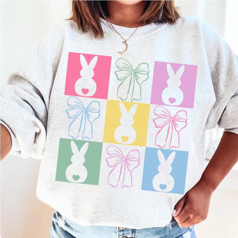 May include: A light gray sweatshirt with a colorful design of pastel-colored bunnies and bows. The design is in a grid pattern with each bunny and bow in a different color square.