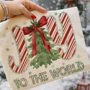 Puede incluir: Un bolso tote navide&ntilde;o color crema con las palabras "JOY TO THE WORLD". La palabra "JOY" est&aacute; en rayas rojas y blancas de bast&oacute;n de caramelo. Un &aacute;rbol de Navidad verde y un lazo rojo est&aacute;n en el centro.