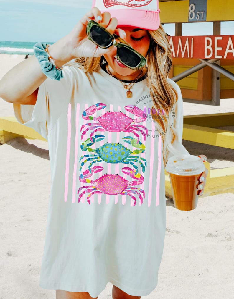 Puede incluir: Camiseta color crema con un dise&ntilde;o de tres cangrejos coloridos apilados verticalmente. Los cangrejos son rosas, azules y rosas, con rayas verticales rosas. La camiseta tiene el texto "TRAINING ORIGINAL" impreso.