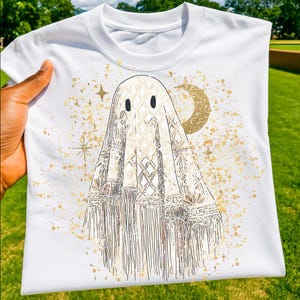 May include: White crew neck t-shirt featuring a ghost design. The ghost is depicted with a macrame-style design, gold accents, and a crescent moon. The design is surrounded by gold stars and speckles.