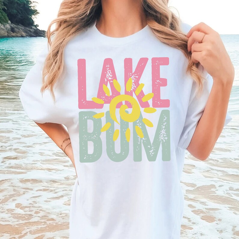 Op de afbeelding: Wit T-shirt met een grafisch ontwerp dat "LAKE" in roze letters en "BUM" in groene letters zegt met een gele zon tussen de woorden.