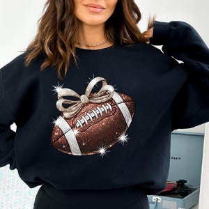Pode incluir: Camisola de moletom preta com uma bola de futebol marrom e branca brilhante com um la&ccedil;o dourado no topo. A bola de futebol &eacute; decorada com strass.