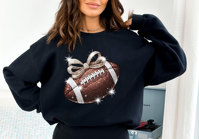 Pode incluir: Camisola de moletom preta com uma bola de futebol marrom e branca brilhante com um la&ccedil;o dourado no topo. A bola de futebol &eacute; decorada com strass.