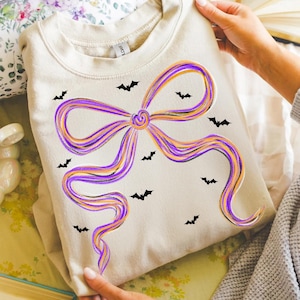 Op de afbeelding: Een crèmekleurige sweatshirt met een groot strikdesign in paars en oranje, met zwarte vleermuissilhouetten. De sweatshirt wordt door een persoon vastgehouden.