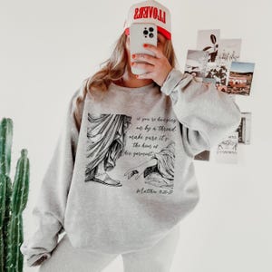 May include: Gray sweatshirt with a black and white illustration of two hands reaching for each other. The text "if you're hanging on by a thread make sure it's the hem of his garment" and "Matthew 9:20-27" are printed on the sweatshirt.