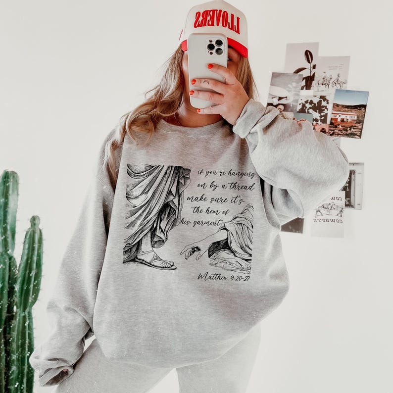 May include: Gray sweatshirt with a black and white illustration of two hands reaching for each other. The text "if you're hanging on by a thread make sure it's the hem of his garment" and "Matthew 9:20-27" are printed on the sweatshirt.