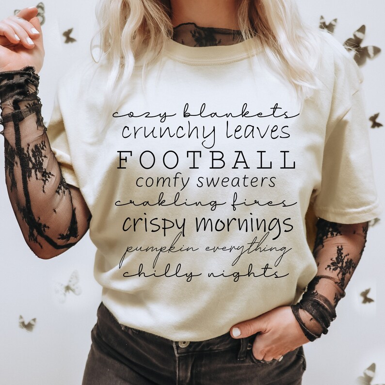 May include: Cream-colored t-shirt with black text listing autumn-themed phrases: "cozy blankets," "crunchy leaves," "FOOTBALL," "comfy sweaters," "crackling fires," "crispy mornings," "pumpkin everything," and "chilly nights."