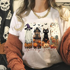 May include: White t-shirt featuring a Halloween-themed graphic. The design includes a ghost wearing a purple witch hat, a haunted house, and a black cat, all surrounded by pumpkins and bats. The words "Spooky Season" are written below the images.