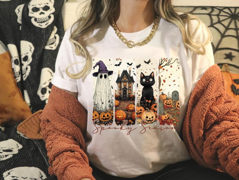 May include: White t-shirt featuring a Halloween-themed graphic. The design includes a ghost wearing a purple witch hat, a haunted house, and a black cat, all surrounded by pumpkins and bats. The words "Spooky Season" are written below the images.