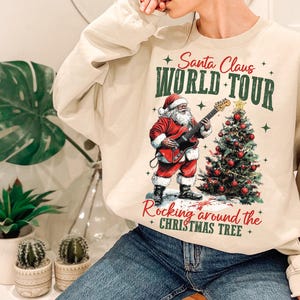 May include: Beige sweatshirt with a graphic of Santa Claus playing a guitar in front of a Christmas tree. Text on the sweatshirt reads "Santa Claus World Tour" and "Rocking around the Christmas Tree."