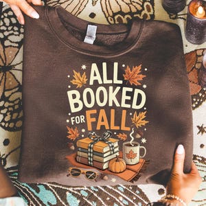 May include: A brown sweatshirt with the text "ALL BOOKED FOR FALL" in white and orange. The graphic includes a stack of books, a coffee mug, a pumpkin, and fall leaves. Sunglasses and acorns are also featured in the design.