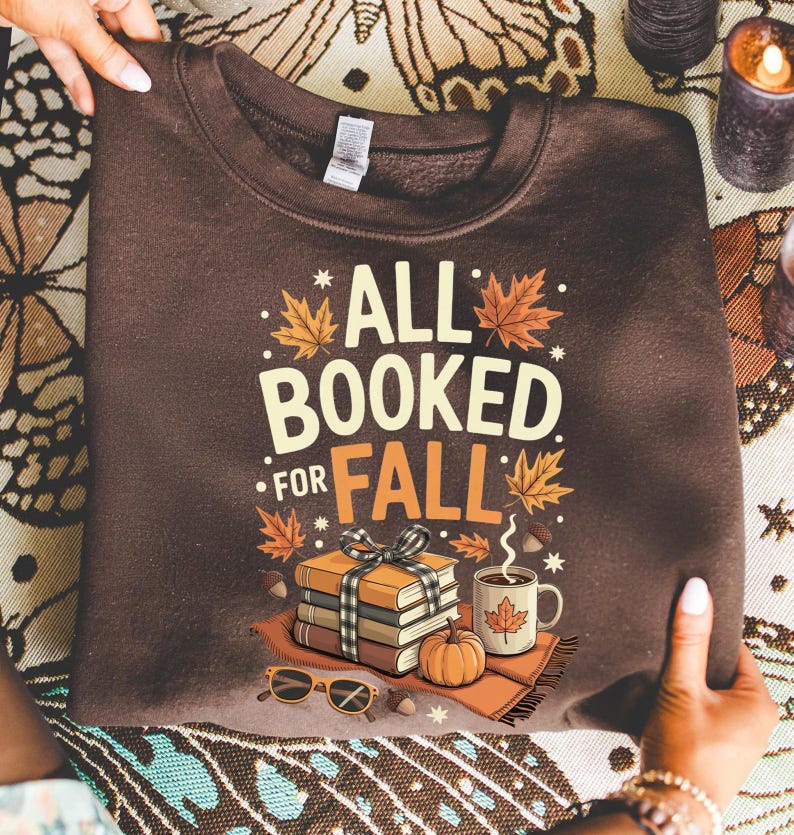 May include: A brown sweatshirt with the text "ALL BOOKED FOR FALL" in white and orange. The graphic includes a stack of books, a coffee mug, a pumpkin, and fall leaves. Sunglasses and acorns are also featured in the design.