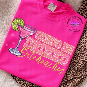 May include: A bright pink t-shirt featuring a margarita glass with a lime wedge and the text "Cinco De Drinko Bitchoachos" in a playful font. The shirt has a crew neck and short sleeves.