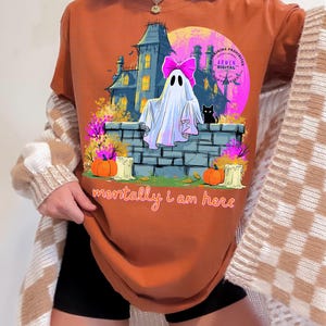 Puede incluir: Una camiseta de color naranja quemado con un estampado gr&aacute;fico de un fantasma con un lazo rosa, un gato negro, una casa encantada, calabazas y velas. El texto "mentally i am here" est&aacute; impreso en letra cursiva naranja en la parte inferior.