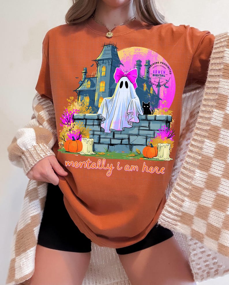 Puede incluir: Una camiseta de color naranja quemado con un estampado gr&aacute;fico de un fantasma con un lazo rosa, un gato negro, una casa encantada, calabazas y velas. El texto "mentally i am here" est&aacute; impreso en letra cursiva naranja en la parte inferior.