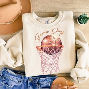 May include: A cream-colored sweatshirt with the text "Game Day" in gold script. A glittery basketball is shown going through a pink and white basketball hoop.