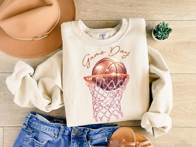May include: A cream-colored sweatshirt with the text "Game Day" in gold script. A glittery basketball is shown going through a pink and white basketball hoop.