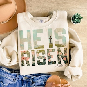 May include: A cream-colored sweatshirt with the words "He is Risen" in large, stylized letters. The letters are filled with a watercolor-style floral design. The text "Matthew 28:6" is written in a smaller font below the main text.