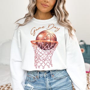 May include: White sweatshirt with a pink and gold glitter basketball in a hoop. The text "Game Day" is written in gold glitter above the basketball.