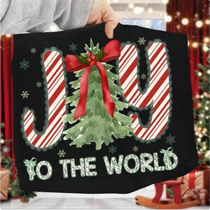 Puede incluir: Coj&iacute;n decorativo negro con tem&aacute;tica navide&ntilde;a con la frase "JOY TO THE WORLD". La palabra "JOY" est&aacute; en rayas rojas y blancas de bast&oacute;n de caramelo con un ribete de encaje verde. Un &aacute;rbol de Navidad de acuarela con un lazo rojo est&aacute; en el centro.