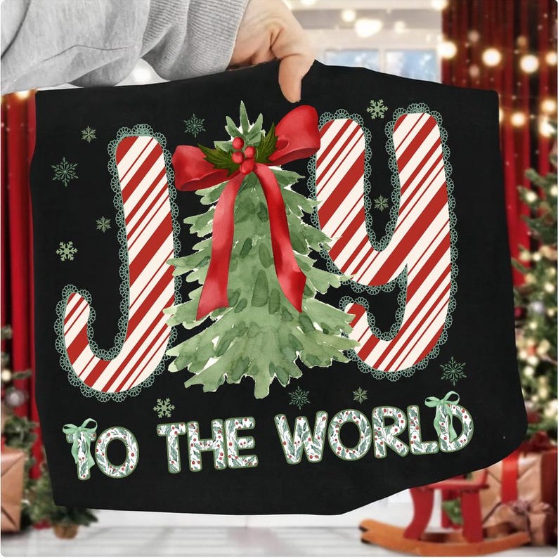 Puede incluir: Coj&iacute;n decorativo negro con tem&aacute;tica navide&ntilde;a con la frase "JOY TO THE WORLD". La palabra "JOY" est&aacute; en rayas rojas y blancas de bast&oacute;n de caramelo con un ribete de encaje verde. Un &aacute;rbol de Navidad de acuarela con un lazo rojo est&aacute; en el centro.