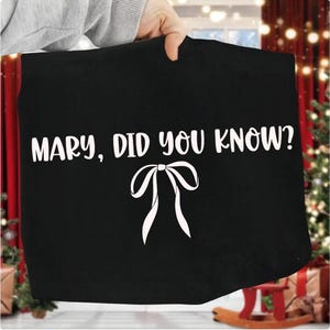 Może przedstawiać: Czarna torba z białym napisem "MARY, DID YOU KNOW?" i białą grafiką kokardy. Torba jest trzymana przez dłoń. Tło zawiera dekoracje świąteczne.