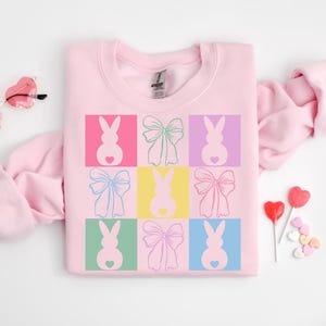 May include: Pink sweatshirt with a graphic design of pastel colored bunnies with bows. The bunnies are in a grid pattern with each bunny in a different colored square.