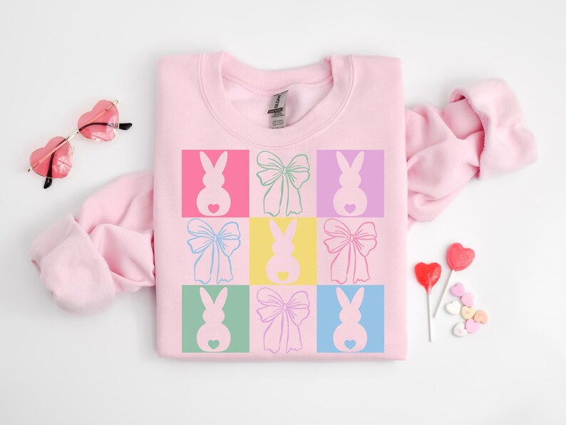 May include: Pink sweatshirt with a graphic design of pastel colored bunnies with bows. The bunnies are in a grid pattern with each bunny in a different colored square.