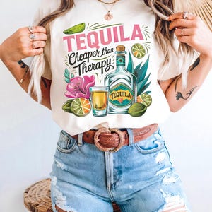 Pu&ograve; includere: Maglietta bianca con un design grafico di una bottiglia di tequila, un bicchierino, lime e fiori. Il testo sulla maglietta recita "Tequila Cheaper than Therapy".