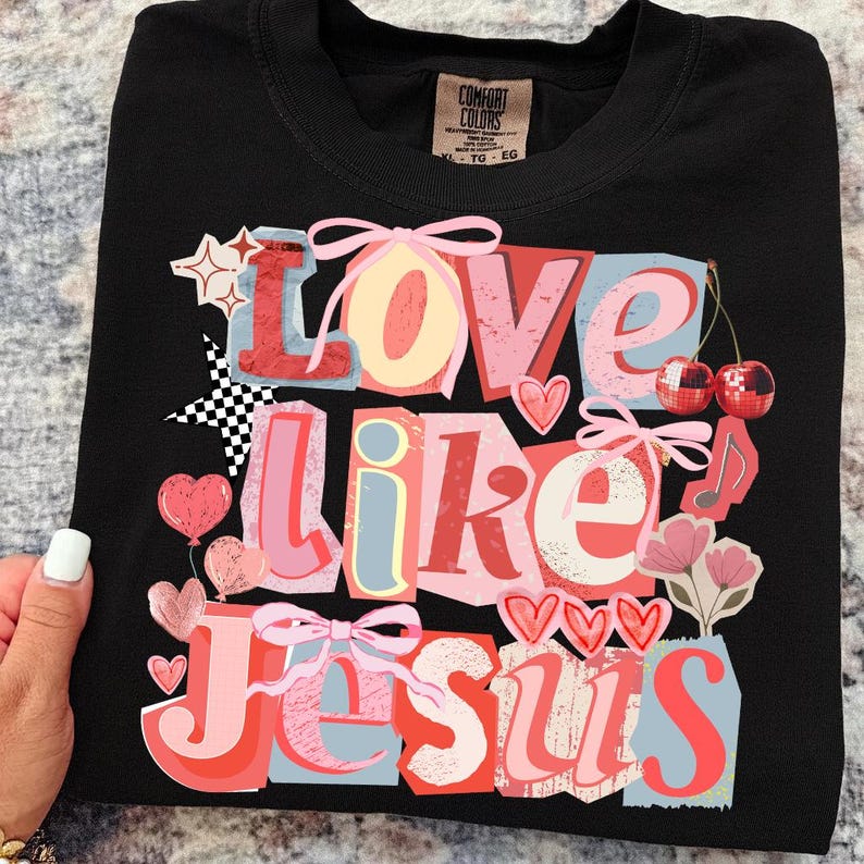 May include: Black t-shirt featuring the words "Love Like Jesus" in a layered, colourful design. The design incorporates hearts, cherries, stars, and bows. The shirt has a crew neck and a "Comfort Colors" label.