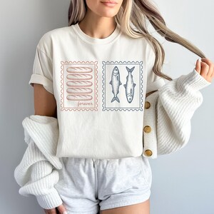 May include: Cream-colored t-shirt featuring two postage stamp designs. One stamp shows five bread loaves, the other two fish. The word "forever" is printed on the bread stamp. The shirt is a casual, everyday wear item.