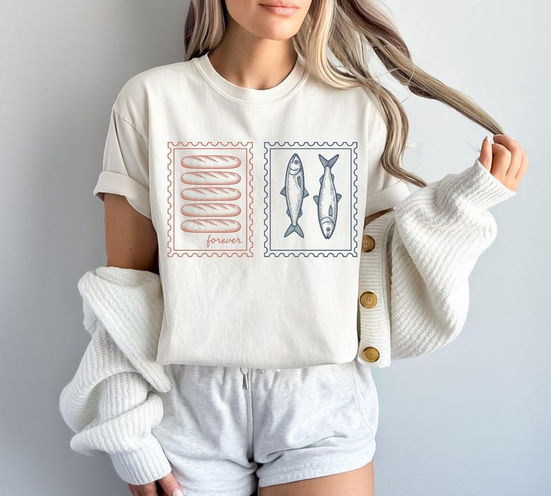 May include: Cream-colored t-shirt featuring two postage stamp designs. One stamp shows five bread loaves, the other two fish. The word "forever" is printed on the bread stamp. The shirt is a casual, everyday wear item.