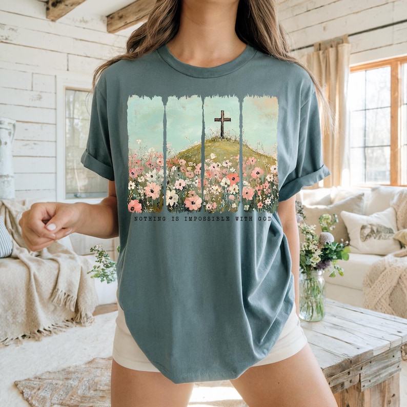 May include: A teal blue t-shirt with a graphic of a wooden cross on a hill with flowers. The text "Nothing is impossible with God" is printed below the graphic.