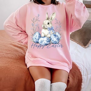 May include: Pink Easter-themed sweatshirt featuring a white bunny with a blue bow, sitting among blue patterned eggs and floral accents. The words "Happy Easter" are written in blue script. The sweatshirt is a long-sleeved, crew neck style.