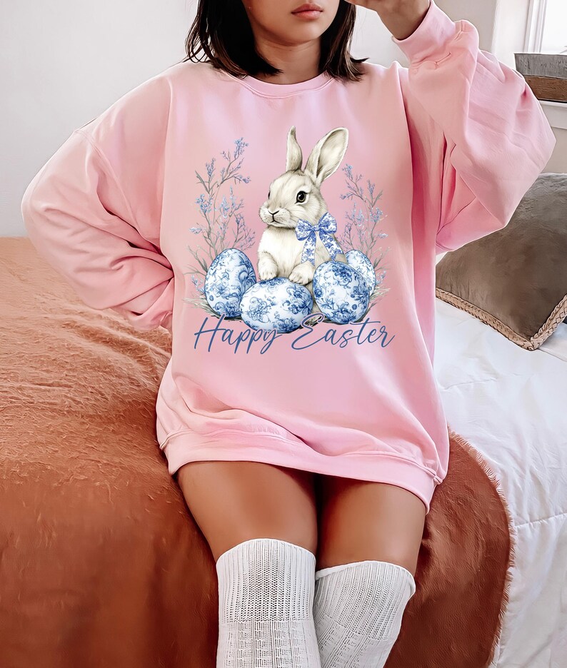 May include: Pink Easter-themed sweatshirt featuring a white bunny with a blue bow, sitting among blue patterned eggs and floral accents. The words "Happy Easter" are written in blue script. The sweatshirt is a long-sleeved, crew neck style.