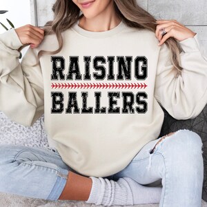 May include: A cream colored sweatshirt with the text "RAISING BALLERS" in black with a red baseball stitch design.