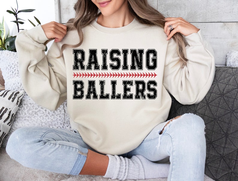 May include: A cream colored sweatshirt with the text "RAISING BALLERS" in black with a red baseball stitch design.