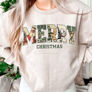 May include: Beige sweatshirt with the words "MERRY CHRISTMAS" in a green, vintage-style font. The letters are filled with Christmas-themed illustrations, including Santa Claus, a gingerbread man, a snowman, and a reindeer.
