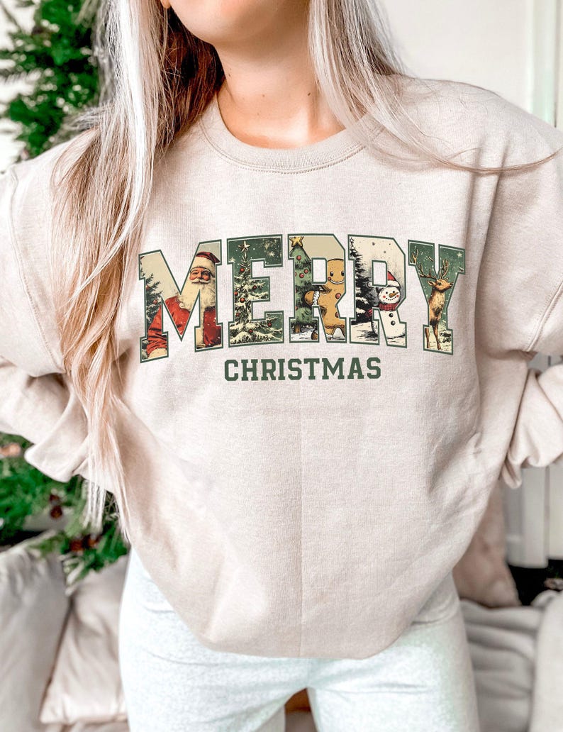May include: Beige sweatshirt with the words "MERRY CHRISTMAS" in a green, vintage-style font. The letters are filled with Christmas-themed illustrations, including Santa Claus, a gingerbread man, a snowman, and a reindeer.